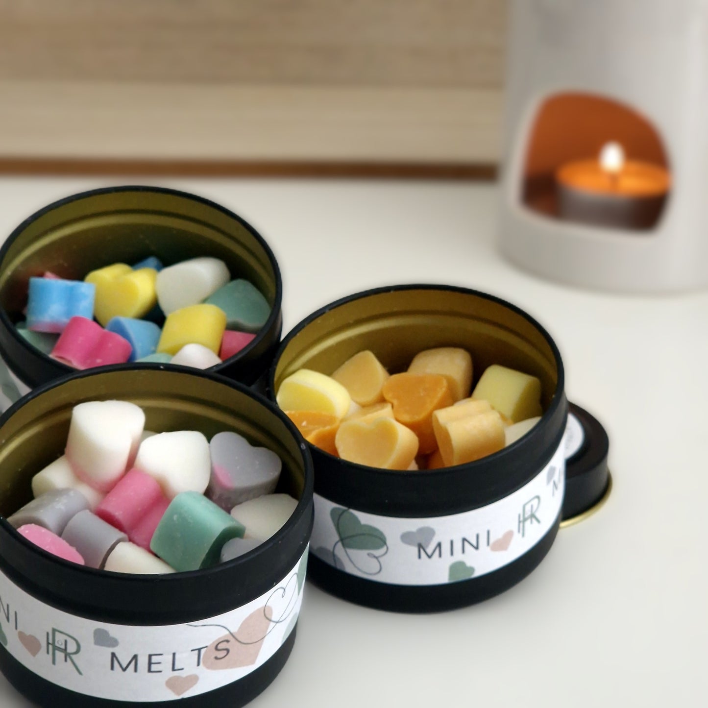 3 small tins of colourful mini heart shaped wax melts with a wax melter in the background 